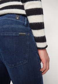 Jeans din denim albastru închis, cu un design ajustat, având buzunare la spate și o etichetă mică cu text. Purtați cu un pulover cu dungi.