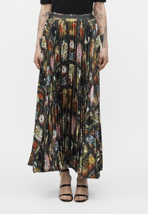 Femme portant une jupe longue plissée avec un imprimé collage coloré, un haut noir et des sandales noires à talons hauts à brides, debout devant un fond uni.