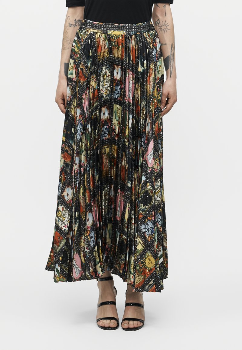 Kobieta ubrana w plisowaną spódnicę maxi z kolorowym nadrukiem kolażu, czarny top i czarne sandały na wysokim obcasie z paskami, stojąca na jednolitym tle.