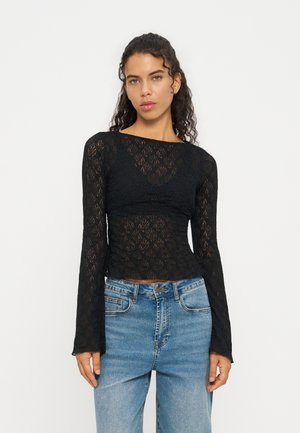 Mujer con cabello rizado que lleva una blusa negra de encaje de manga larga y vaqueros azules, de pie frente a un fondo blanco liso.