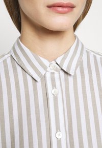 Chemise rayée présentant des rayures horizontales blanches et beige clair, un col pointu et des boutons blancs sur le devant. Tissu lisse et texturé.