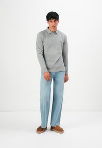 Męski model stoi w szarym longsleeve polo, jasnoniebieskich szerokich dżinsach oraz brązowych butach wsuwanych na tle jednolitego białego tła.