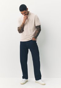 Ljusbeige pikétröja med korta ärmar, marinblå jeans och vita sneakers. Piket har en diskret logga på bröstet. Byxorna har en avslappnad passform.