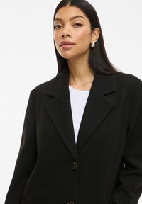 Cappotto in misto lana nero con colletto a punte, due bottoni e tasche laterali. Indossato sopra un top bianco, con tessuto liscio e testurizzato.