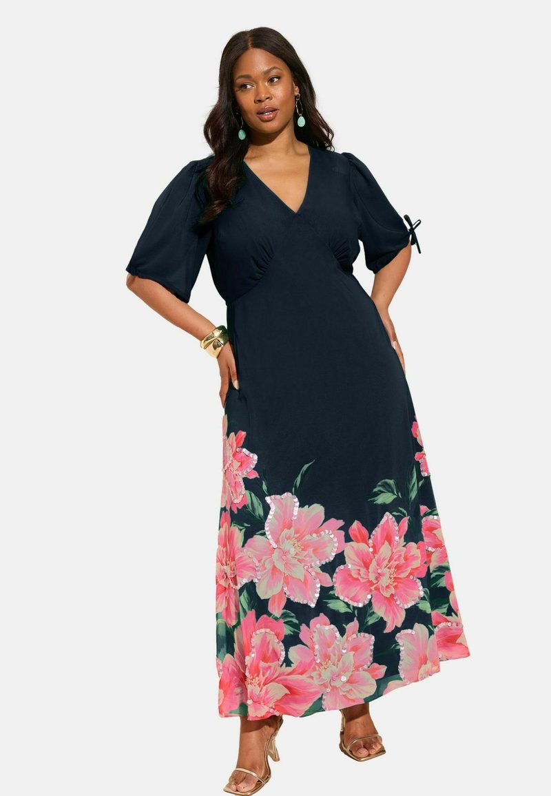 Naine, kes kannab tumesinist maksikleit suurt roosat lillemustriga allosas, lõhitatud varrukatega, millel on paelad, ning kuldseid kontsakingi.