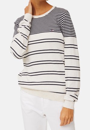 Donna che indossa un maglione bianco a maniche lunghe con righe orizzontali nere, abbinato a pantaloni bianchi, mano che tocca il collo, in piedi su sfondo semplice.