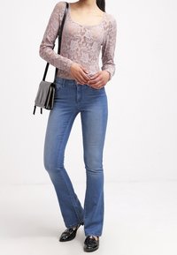 Top de encaje de manga larga en rosa claro, combinado con jeans acampanados de talle medio en azul claro. Un bolso negro y zapatos negros completan el conjunto.