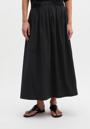 Femme portant une longue jupe plissée noire et des sandales noires ouvertes, debout sur un fond blanc.