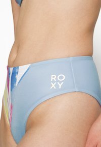 Parte de baixo de fato de banho azul claro com um padrão abstrato colorido e o logotipo dourado "ROXY". Textura suave e design de cintura alta com cobertura moderada.