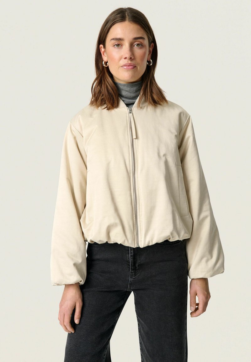 Beige bomberjack met een ritssluiting aan de voorkant, elastische zoom en ronde kraag. Beschikt over ontspannen mouwen en een zachte textuur. Gepaard met zwarte jeans.