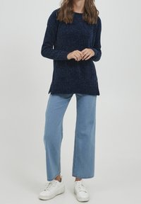 Femme portant un pull en maille bleu marine, un jean large bleu clair et des baskets blanches, debout les mains légèrement croisées.