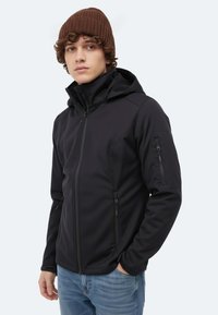 Schwarze Softshell-Jacke mit hohem Kragen und verstellbarer Kapuze. Verfügt über Reißverschlusstaschen und eine glatte, strukturierte Oberfläche. Kombiniert mit blauer Jeans.