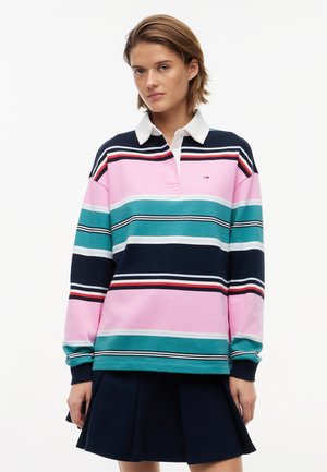 Kvinde iført langærmet rugbytrøje med horisontale striber i pink, teal, navy og hvid, kombineret med en navy plisseret nederdel.