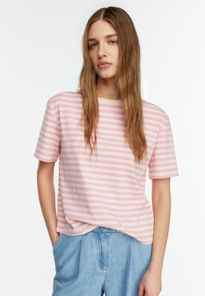 Junge Frau mit langem hellbraunem Haar, die ein kurzärmeliges Shirt mit rosa-weißen Streifen und hellblaue Jeans trägt, steht mit einer Hand in der Tasche.