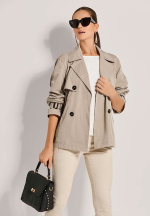 Beige dubbelzijdige trenchcoat met grote zwarte knopen, textuur witte top, beige broek en een zwarte handtas met gouden accenten.