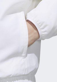 Hvid letvægtsjakke med elastiske manchetter og sidelomme. Glat tekstur med et subtilt rutemønster. Enkel, minimalistisk design.