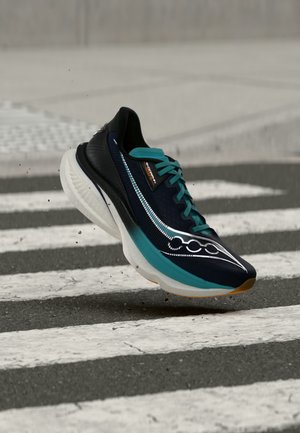 Une chaussure de course noire et turquoise, dotée d’une épaisse semelle blanche, flotte au-dessus d’une piste rayée, avec des gouttelettes éparpillées autour.