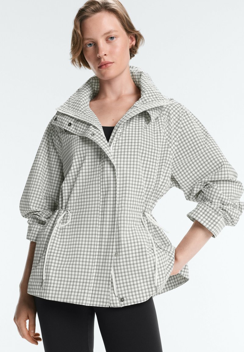 Femme portant une veste à carreaux claire avec des cordons à la taille et des manches retroussées, debout sur un fond uni.