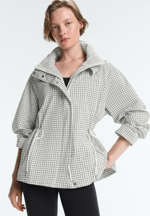 Frau trägt eine hell karierte Jacke mit Kordelzug in der Taille und aufgerollten Ärmeln, steht vor einem einfarbigen Hintergrund.