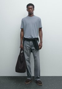 Šedé tričko s krátkým rukávem, světle šedé džíny, tmavá semišová taška a hnědé boty. Jednoduchý design s nabíraným svetr na pase.