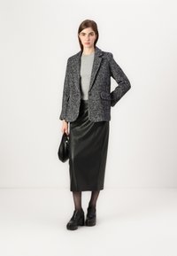 Blazer en chevrons noir, pull à côtes gris, jupe midi en cuir noir, talons épais noirs et un sac à main noir sur un fond blanc.