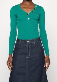 Mujer vistiendo un top de canalé en color verde azulado de manga larga con botones dorados cerca del escote en V y una falda de mezclilla oscura de cintura alta.