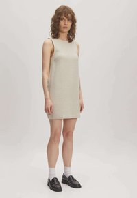 Beige mouwloos jurkje met een rechte snit en een textuurstof. Model draagt zwarte loafers en witte sokken. Eenvoudig en minimalistisch ontwerp.
