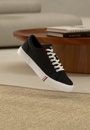 Zwarte lage sneaker met witte veters en zool, rood- en blauwgestreepte details op de zool, zwevend boven een beige tapijt in een woonkameromgeving.