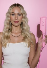 Planchador de pelo rosa con acentos estriados sostenido en la mano, blusa blanca texturizada, cabello rubio ondulado, frente a un fondo rosa.