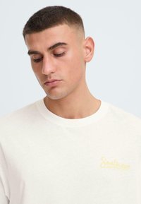 T-shirt en coton blanc à col rond avec un petit logo jaune "Soulscape" sur le côté gauche, texture lisse et coupe décontractée.