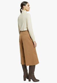 Maglione beige a collo alto abbinato a pantaloni marroni a vita alta e gamba larga. Il modello indossa stivali marroni alti al ginocchio con tacco a blocco.