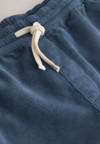 Primo piano di pantaloni di velluto a coste blu con un cordino beige annodato in vita elastica.