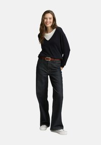 Glunderende vrouw staat met haar handen in haar zakken, draagt een zwarte V-hals trui over een witte top, donkere wijde jeans, een bruine riem en witte sneakers.
