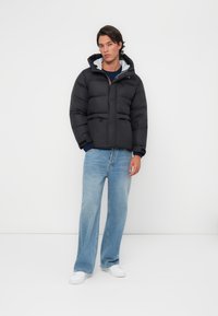 Doudoune noire avec coupe décontractée, fermeture éclair et capuche. Associée à un pantalon en denim large bleu clair et des baskets blanches, sur fond uni.