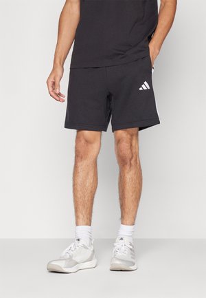 Homme portant un short Adidas noir, des chaussettes blanches et des baskets blanches Adidas, debout devant un fond uni.