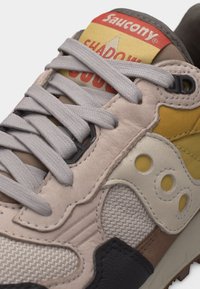 Κοντινό πλάνο αθλητικού παπουτσιού Saucony Shadow σε μπεζ και κίτρινο, με γκρι κορδόνια και κόκκινο λογότυπο στη γλώσσα.