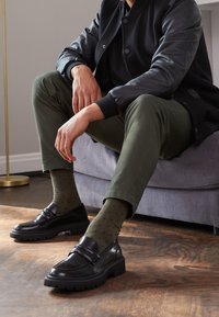 Homme assis sur un canapé gris portant une veste noire, un pantalon vert olive, des chaussettes olive à pois bleu marine, et des mocassins en cuir noir sur un sol en bois.