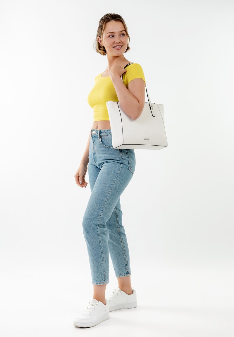 Sac fourre-tout blanc avec des accents noirs, design structuré et double poignées. Modèle portant un crop top jaune, un jean bleu clair et des baskets blanches.