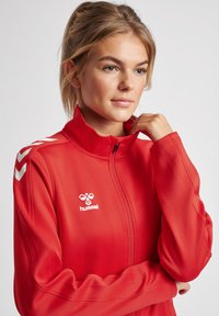 Rode jacket met rits en hoge kraag, lange mouwen, en witte logo- en chevronaccenten op de schouders. Glad stoftextuur.