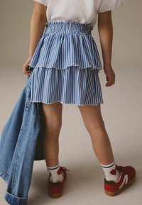 Jupe à volants rayée bleu et blanche avec des bords effilochés, portée avec des chaussures rouges et des chaussettes blanches à rayures bleues, maintenue avec une veste en denim.