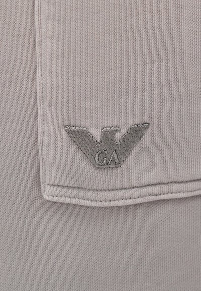 Ανοιχτό γκρι ύφασμα με ριγωτή υφή διαθέτει ένα γκρι κεντημένο λογότυπο ενός αετού με το "GA" από κάτω. Είναι ορατή η προσεγμένη λεπτομέρεια ραφής.