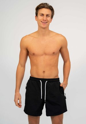 Lachende shirtloze man in zwarte zwembroek met witte trekkoorden, staand tegen een effen lichtgrijze achtergrond.