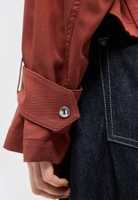 Κοντινή λήψη μανσετών πουκαμίσου σε σκουριά χρώμα με ασημένιο κουμπί και τσέπης σκούρου τζιν με ορατό ράψιμο.