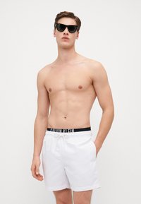 Bela kupaćica s crnim elastičnim pojasom na kojem je istaknut logo "CALVIN KLEIN", glatke teksture i bočnim džepovima. Model nosi crne sunčane naočale.