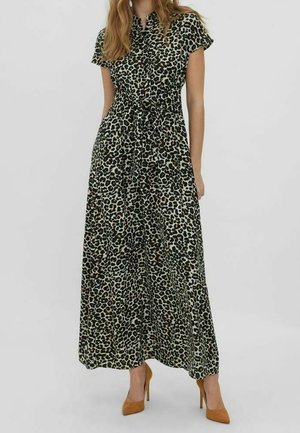 Kobieta ubrana w maxi sukienkę w panterkę z krótkim rękawem i guzikami, z paskiem w talii, w połączeniu z beżowymi wysokimi obcasami, stojąca na białym tle.