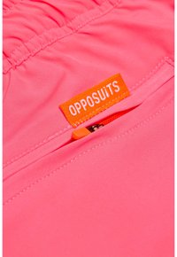 Rožinė medžiaga su susiraukšlėjimo dizainu ir oranžiniu užtrauktuko kišeniu. Yra etiketė su užrašu "OPPOSUITS" ryškiu baltu šriftu ant oranžinio fono.