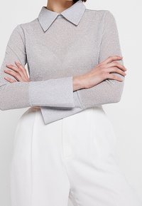 Blouse longue à manches longues en voile argenté avec une finition texturée, dotée d'un col classique et de larges poignets, associée à un pantalon blanc sur mesure.
