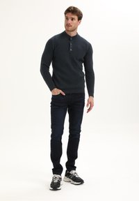 Jongen die met één hand in zijn zak staat, gekleed in een donkere knooptrui, donkere jeans en zwart-witte sneakers.