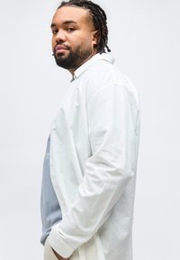 Άνδρας με πλεξούδες στα μαλλιά και γενειάδα, φοράει λευκό πουκάμισο πάνω από ένα μπλε κοντομάνικο με γραφικά, στέκεται πλαγίως με τα χέρια στις τσέπες, μπροστά σε λευκό φόντο.