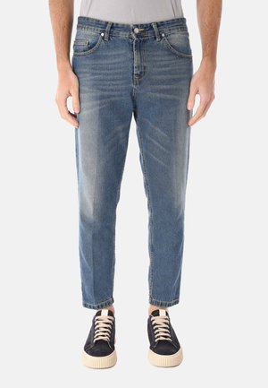 Uomo che indossa jeans a gamba dritta blu sbiaditi e sneakers basse nere con lacci bianchi, in piedi contro uno sfondo chiaro e uniforme.